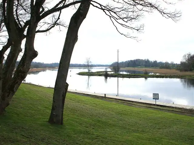 Ein langer Steg zieht sich am Fuß einer Rasenböschung am River Erne bei Killyhevlin entlang, eingerahmt von kahlen Bäumen im Vorfrühling.