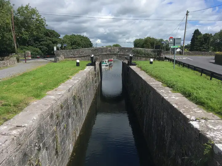 A narrow stone lock chamber full of dark water leads to the arch of Thomastown Bridge, under which two moored cruisers can be seen, with grassy banks and a quiet road on either side.