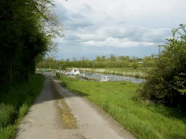 Een pad loopt naar de ligplaatsen van Aghalane aan de Woodford River, met boten aan de steiger en vlakke groene weilanden daarachter.