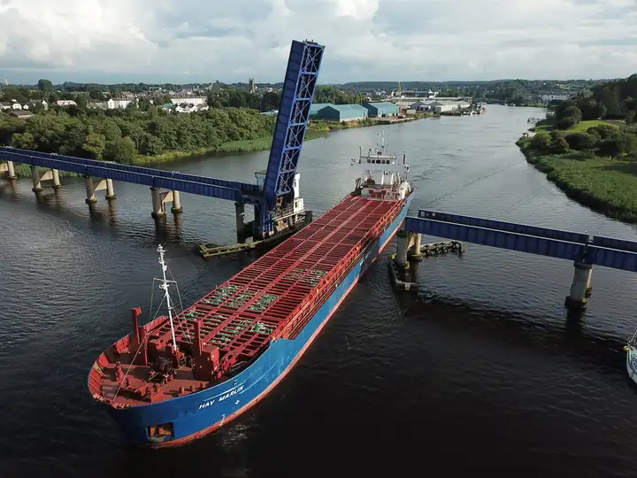 Aerial view of a blue-hulled cargo ship passing through the raised lifting span of the railway bridge over the Bann at Coleraine, with the town and its quays beyond.