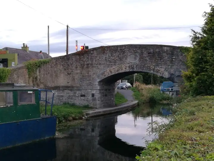 The stone arch of Hazelhatch Bridge reflected in the canal, with a green-and-blue boat moored in the foreground and a covered cruiser under the far bank.