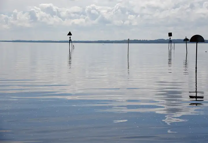 Mehrere Fahrwasserzeichen auf Pfählen stehen im ruhigen, silbrigen Wasser des Lough Neagh vor Toome, am Horizont ein flaches Ufer.