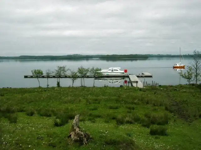 A white cruiser lies at the long timber jetty of Inishmacsaint, lined with young trees, while a small sailing boat is moored offshore on the calm lough.