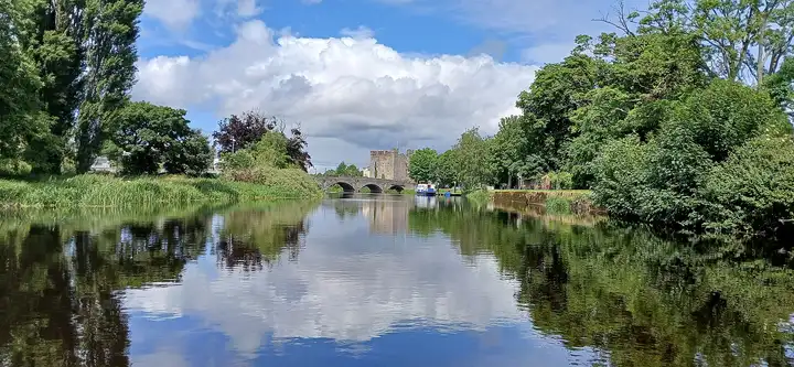 Approaching Athy on the River Barrow, with White’s Castle and the arches of Crom-a-Boo Bridge ahead and clouds reflected in calm water.
