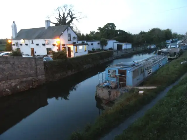 Crépuscule à Hazelhatch, avec des lumières allumées dans un bâtiment blanc au bord du canal et une file de péniches et de bateaux amarrés le long du chemin de halage.