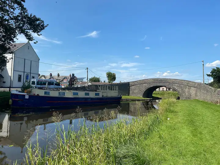 A large blue-and-cream barge moored by Bury Bridge in Tullamore on a sunny day, its hull reflected in the canal beside a grassy towpath.