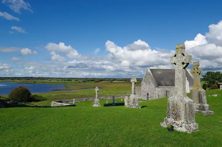Stone crosses and Temple Connor on the grassy slope at Clonmacnoise, above a bend of the Shannon and its flat green callows.