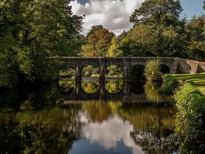 Die Deerpark Bridge, eine alte Steinbrücke mit mehreren Bögen im ehemaligen Schlosspark von Antrim, spiegelt sich zwischen Bäumen im stillen Fluss.