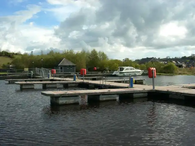 The floating pontoons of Keshcarrigan marina seen from the lough, with red service posts, a single moored cruiser and the houses of the village behind the trees.