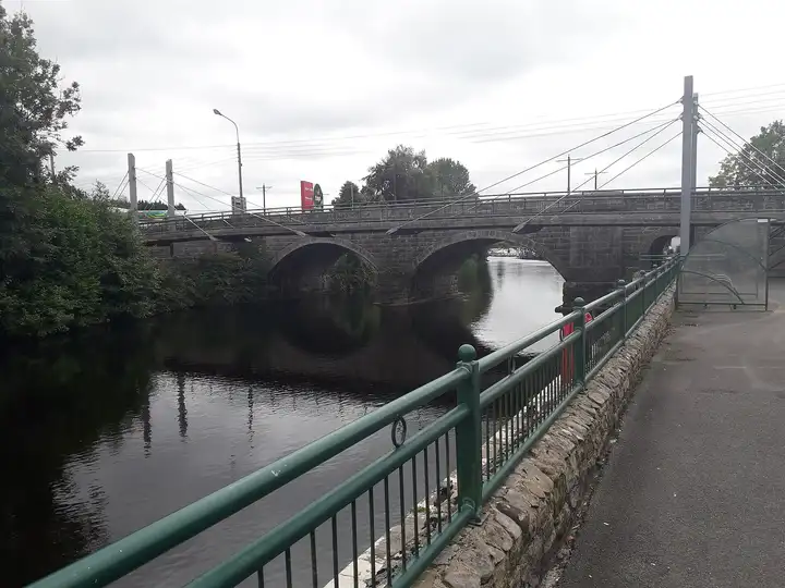 Ballyconnell Bridge, a stone arch bridge over the Woodford River, seen from the railed riverside path, with moored boats visible through one of the arches.