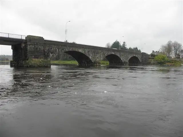 The stone arches of Portglenone Bridge span the grey, fast-flowing Bann, seen from near water level on an overcast day.
