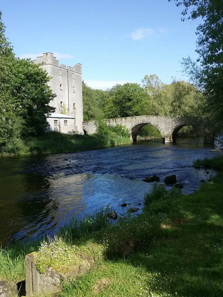 The tall stone mill at Milford beside an arched stone bridge, with the River Barrow running over stones in the foreground.
