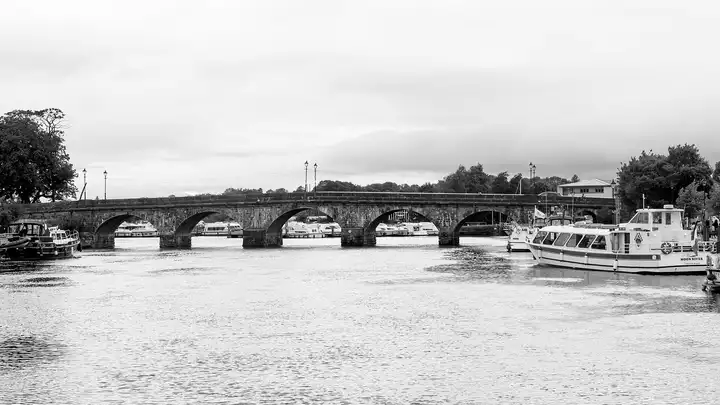 In zwart-wit omlijsten de bogen van de brug van Carrick-on-Shannon rijen afgemeerde kajuitboten, met rechts een rondvaartboot aan de kade.