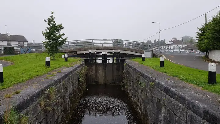 Eine Schleusenkammer an der Cox’s Bridge in Tullamore, die Tore unter der Straßenbrücke mit Geländer geschlossen, auf dem Rasen weiß bemalte Poller.