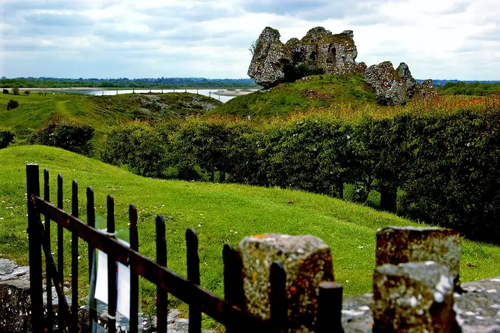The tilted ruins of Clonmacnoise Castle on a grassy mound, with the Shannon winding through the callows behind and an iron gate in the foreground.