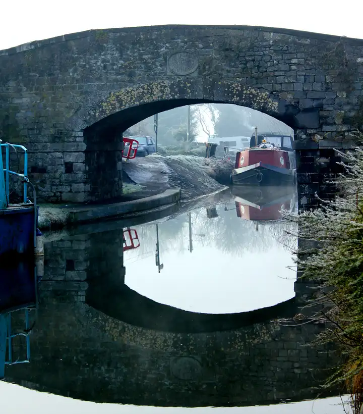 Le pont de Hazelhatch par un jour de givre et de brume, son arche reflétée dans le canal immobile et des bateaux amarrés visibles sous la voûte.