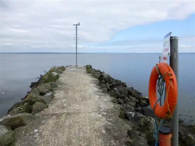 Une jetée en béton bordée de rochers s’avance dans le Lough Neagh à The Battery, avec une bouée de sauvetage et un mât d’éclairage.