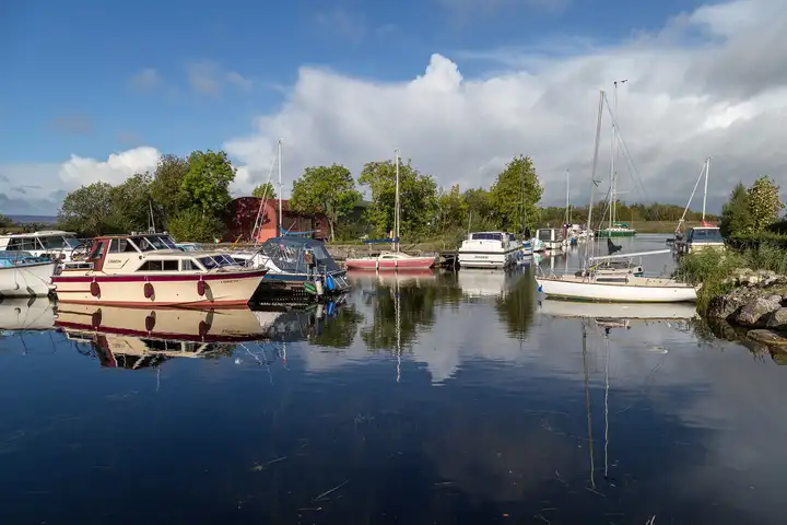 Motorkruisers en zeilboten liggen in de kleine haven van Kilgarvan Quay en spiegelen in het kalme water, met een grote loods en bomen erachter.
