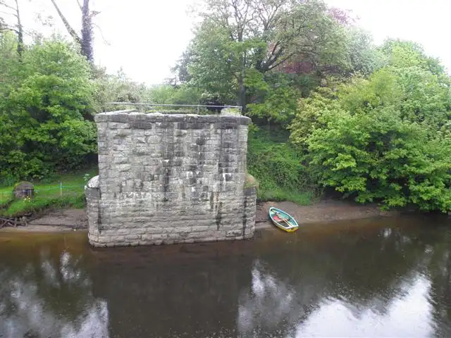 Een losstaande stenen brugpijler aan de rand van de Woodford River bij Aghalane, met een klein roeibootje op de oever ernaast.