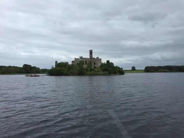 McDermott’s Castle rises above the trees of Castle Island, seen from a boat on Lough Key under a grey sky, with a small rowing boat to the left.