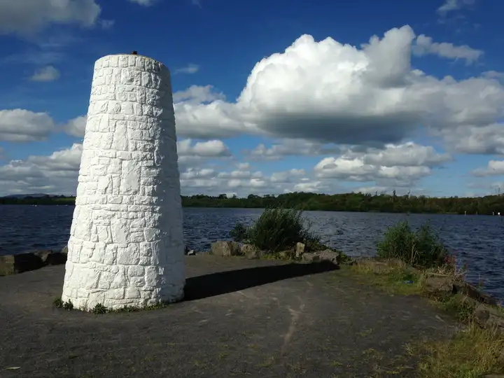 A white-painted stone beacon stands on Rossmore Point at Castle Archdale, with the dark-blue lough and wooded shores under towering white clouds.