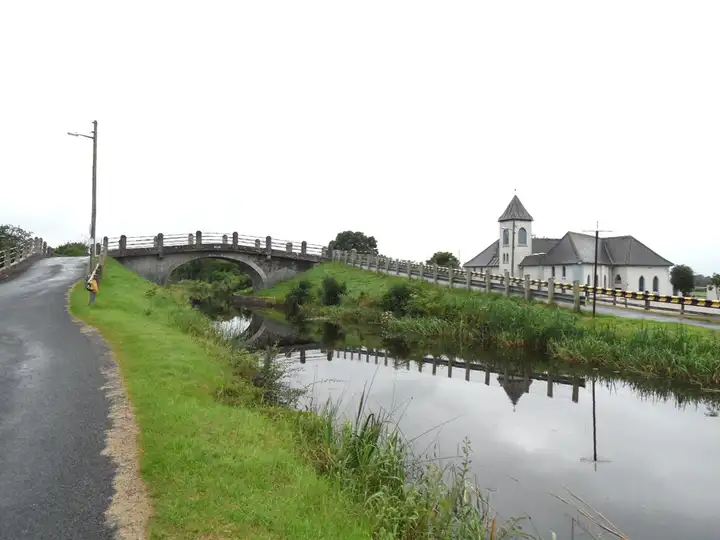 Plunkett Bridge carrying the road over the canal at Pollagh, beside the village church with its small bell tower, the bridge reflected in the water.