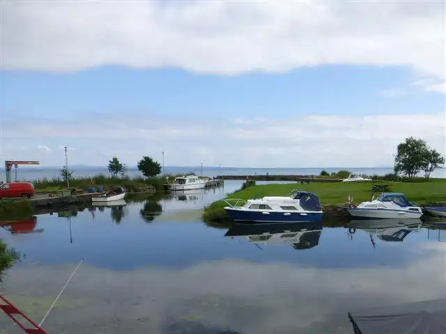 Motor boats moored in the small Battery harbour on Lough Neagh, reflected in calm water, with a jib crane, a jetty and the open lough beyond.