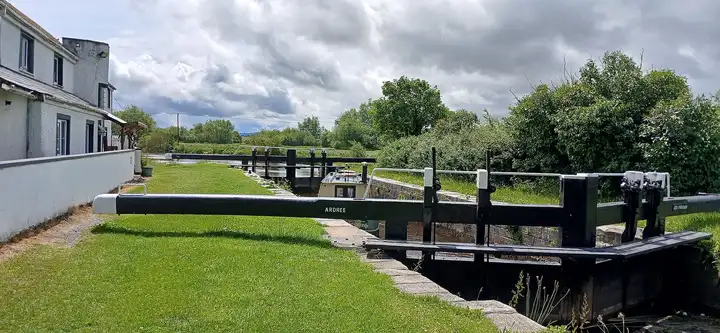 Les balanciers noirs de l’écluse d’Ardreigh sur une pelouse tondue, avec la maison éclusière à gauche sous un ciel nuageux.