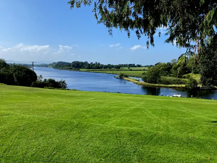 Gemähte Rasenflächen fallen bei Killyhevlin zum River Erne ab, rechts kleine Boote an einem Steg, dahinter windet sich der Fluss in die Ferne.