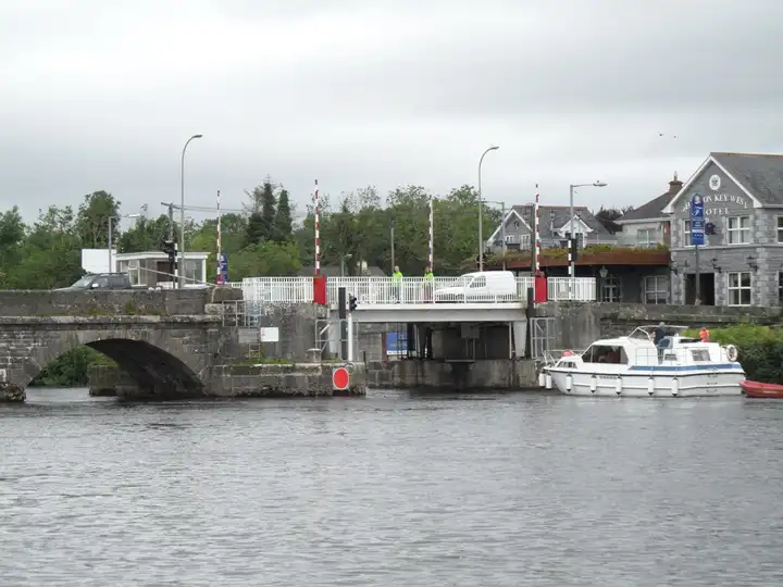 The lifting span of Roosky Bridge, between red-and-white barrier poles, seen from the water beside a stone arch, with a cruiser moored below the hotel on the right.