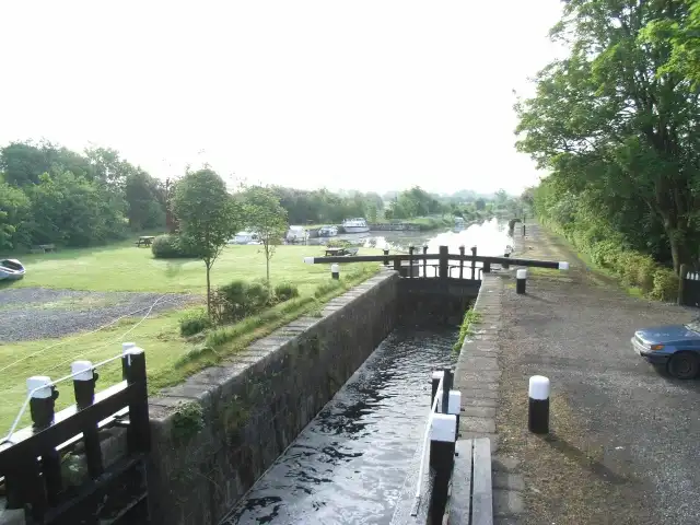 Water rushes through Lock 26 at Coolnahay towards the camera, with the harbour beyond, where boats are moored beside wide lawns and picnic tables.