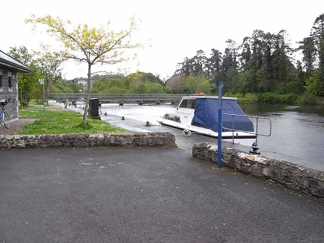 Een kruiser met een blauw dekzeil aangemeerd in Ballinamore, met een voetbrug over de vaarweg en hoge bomen op de andere oever.