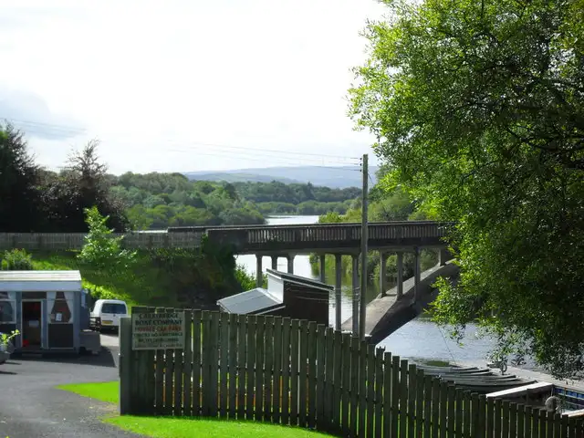 Carry Bridge crosses the Erne channel on its piers beyond a wooden fence and boatyard sheds, with wooded hills in the distance.