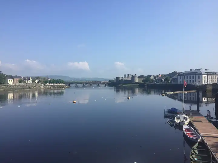 Small boats lie at a pontoon on the calm Shannon in Limerick city centre, with King John’s Castle and Thomond Bridge upriver.