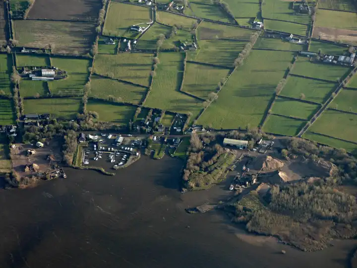 Aerial view of the Lough Neagh shore at Sandy Bay, with boats in and around the small marina basin, sand heaps at a quay to the right and patchwork fields behind.