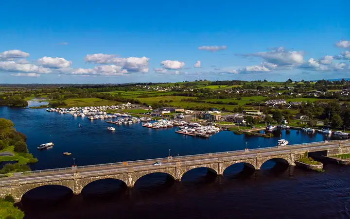 Aerial view of the stone arches of Banagher Bridge over the Shannon, with rows of cruisers in the marina beside it and green countryside beyond.