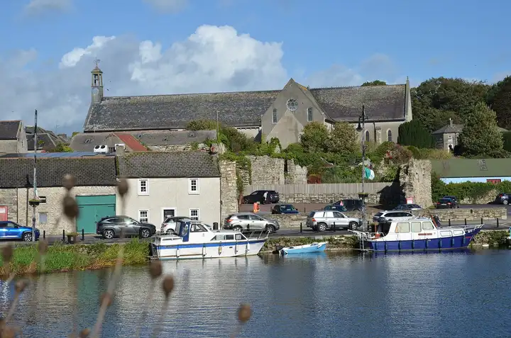 Boats moored at Graiguenamanagh quay below Duiske Abbey church and old stone buildings, seen across the river on a sunny day.