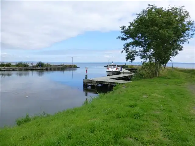 Un appontement en bois avec un bateau de croisière amarré dans le port de The Battery, un arbre feuillu sur la berge herbeuse et le Lough Neagh au-delà de la digue.