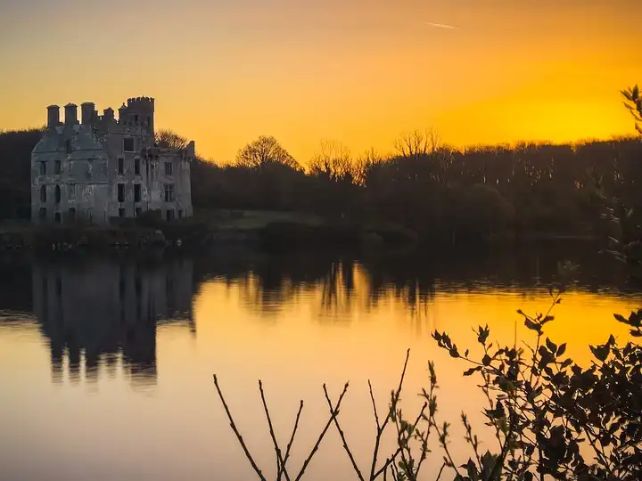 Une large étendue lisse de la rivière Corrib à Menlo à l’aube, avec la ruine du château sur la rive opposée à gauche et un ciel orangé au-dessus des arbres.