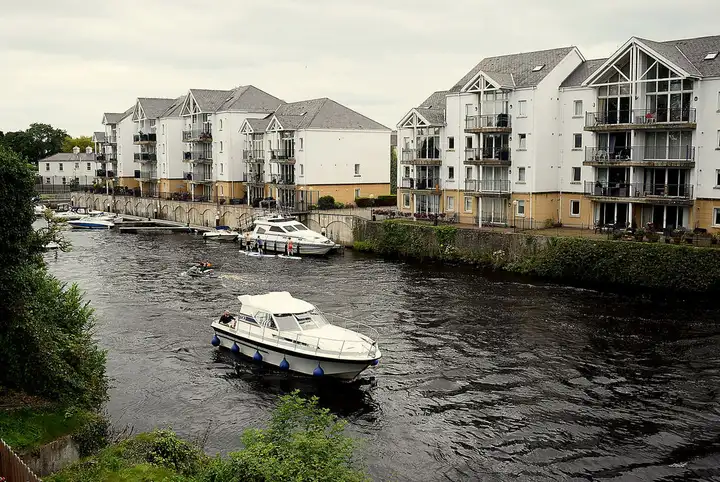 Ein Motorkreuzer fährt in Enniskillen auf dem River Erne an einer Reihe weißer Wohnblocks am Ufer vorbei, am anderen Ufer liegen weitere Boote.