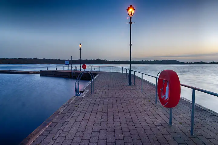 The paved public quay at Lanesborough-Ballyleague at dusk, with lit lamps, railings and a red lifebuoy beside the calm water of the Shannon.