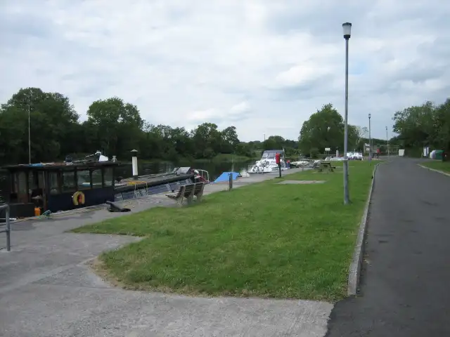 Boats lie along the mooring at Drumsna Harbour, beside a lawn with benches and a lamp-lit road on an overcast summer day.