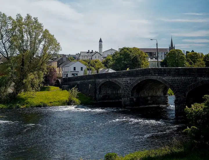 The arched stone Erne Bridge spans the rippling River Erne at Belturbet, with the town’s houses and two church towers rising behind the trees.