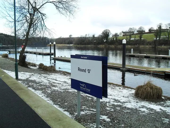 Ein Namensschild des Round O steht auf einem verschneiten Weg neben den Pontons am Round O Quay in Enniskillen, dahinter der River Erne und bewaldete Hügel.