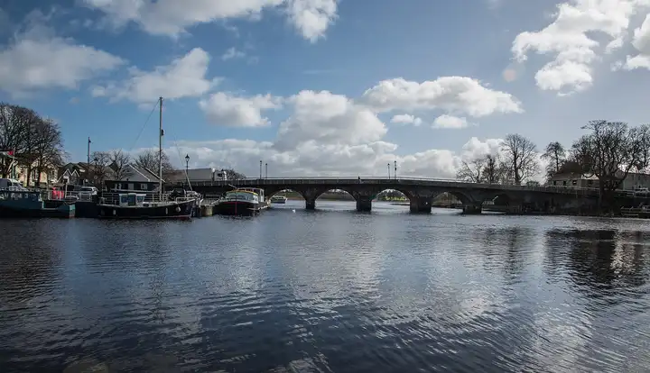 The arched stone bridge at Carrick-on-Shannon spans the wide river under a blue sky with white clouds, with a sailing boat and a barge moored at the quay on the left.