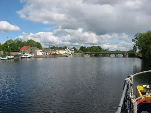 Roosky gezien vanaf een boot op de Shannon, met afgemeerde boten langs de dorpskade en de brug met vele bogen over de rivier onder een bewolkte blauwe lucht.
