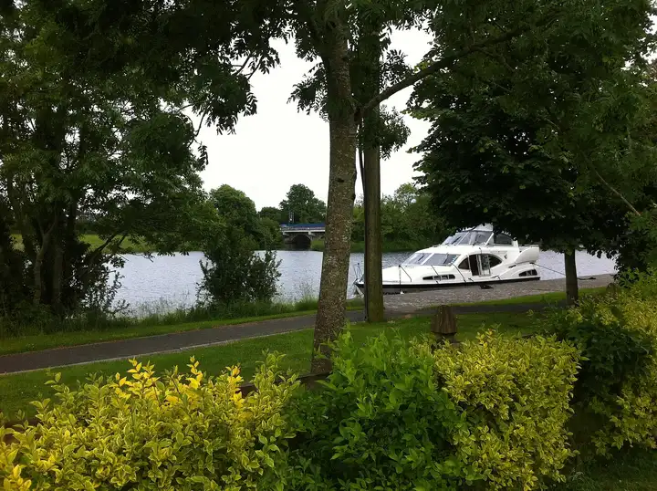 A white cruiser lies moored at the stone quay in Tarmonbarry, seen through trees and shrubs from the riverside path, with the N5 bridge in the distance.