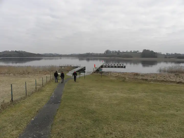 A few visitors follow a path across the grass of Devenish Island to its timber jetties on the calm lough, with low green hills beyond.