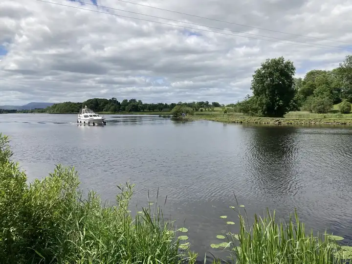 A white motor cruiser heads along the broad River Erne at Killyhevlin, between reedy banks and wooded shores under a cloudy sky.