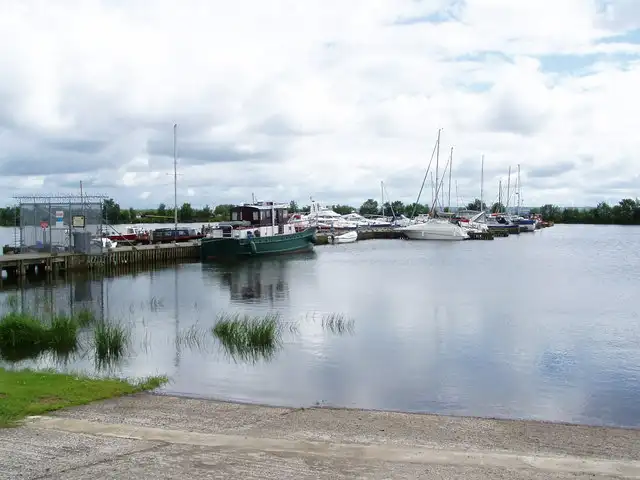 Boats moored along the jetties of Ballyronan Harbour, seen from the slipway across calm water under a cloudy sky.